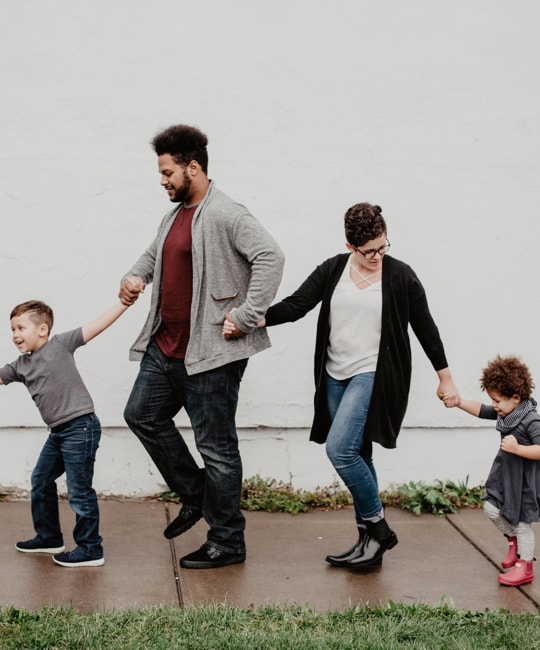 Family of four holding hands while walking together on a wet sidewalk against a white brick wall